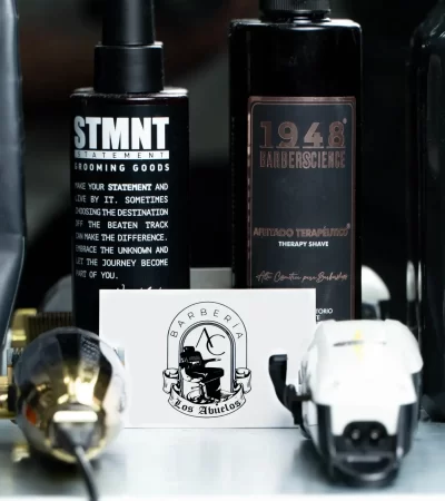 Productos de barbería en una mesa, incluyendo botellas de STMT y 1948 Barberscience, junto a una tarjeta de presentación de "Barbería Los Abuelos" y herramientas de corte. Ideal para promocionar servicios de grooming y afeitado terapéutico.
