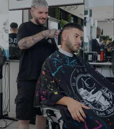 Barbero cortando el cabello de un cliente en una barbería moderna, con un enfoque en el ambiente estilizado y profesional del salón. El cliente lleva una capa con un diseño colorido mientras el barbero utiliza herramientas de corte.