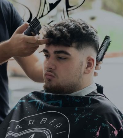 Hombre joven con cabello rizado recibiendo un corte de pelo en una barbería, mientras un barbero utiliza una máquina de cortar y un peine. El cliente lleva una capa de corte con un diseño colorido.